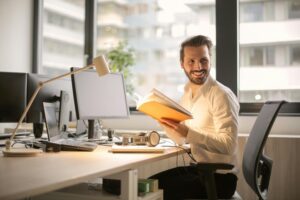 A man sitting at an office desk with documents and a laptop, smiling in a bright workplace setting.